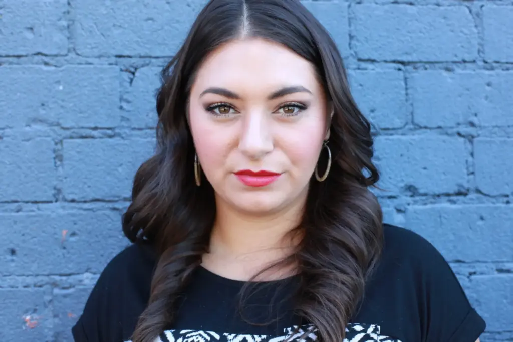 Sonia Gomez with long dark hair and bold red lipstick, standing against a textured blue wall, embodying empowerment and community engagement in discussions on health and sustainability.