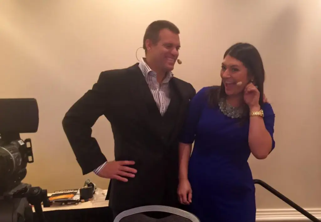 Sonia Gomez and a male colleague posing together at a professional event, both wearing microphones, with a camera and equipment visible in the background.