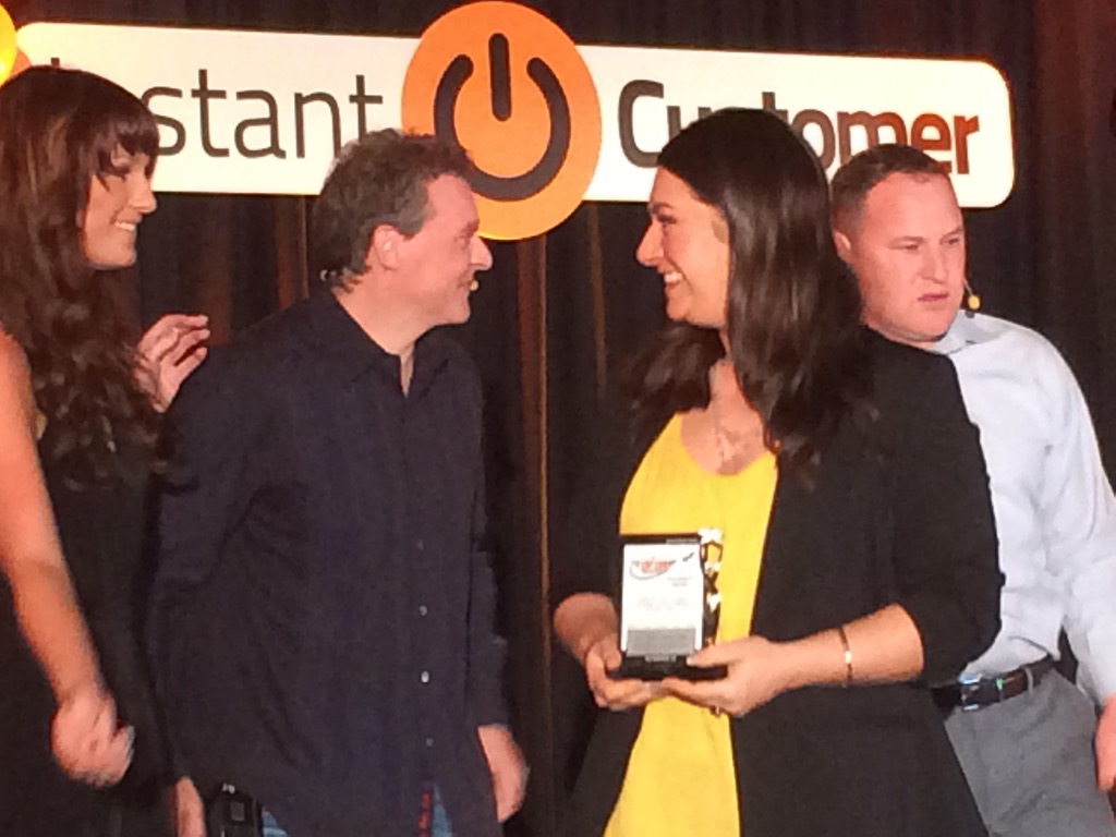 Sonia Gomez receiving an award on stage, surrounded by colleagues, with the "Instant Customer" logo in the background.